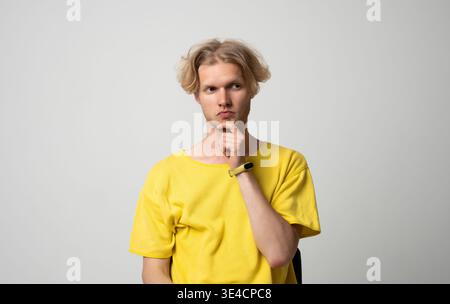 Young man with blond hair in yellow t-shirt pondering a solution. Contemplating complex ideas. Feeling confused and in doubt about making a decision Stock Photo