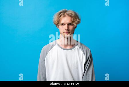 Young casual man with a serious expression and blonde wavy hair in a raglan shirt, standing confidently against a bright blue background, copy space Stock Photo