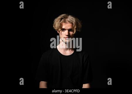 Young blond man with beard looking directly at camera, standing in a studio with a dark background, portraying seriousness and introspection Stock Photo