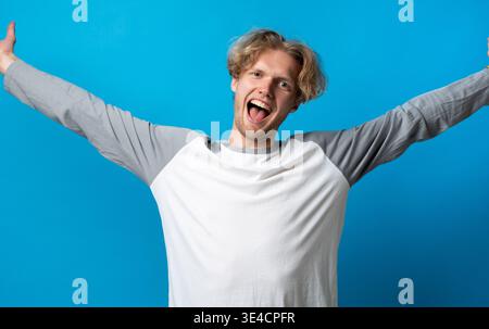 Excited young man with outstretched arms celebrating success, expressing happiness and joy on blue background, feeling triumph and achieving goals Stock Photo