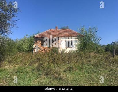 abandoned and broken small house, with red tiles roof, white wall, window and green grass in front of house. Blue sky is above the house. Stock Photo