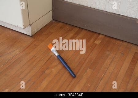Orange brush lies on wooden floor. Blue handle contrasts with warm flooring tones. Corners show baseboards and white wall. Tool rests casually, unused Stock Photo