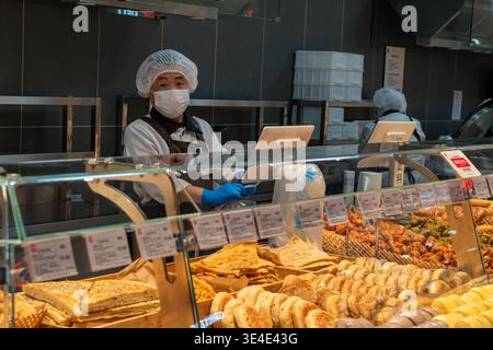 Beijing, China - March 27 2026: Masked male supermarket worker in uniform behind a glass deli counter filled with traditional Chinese baked goods. Stock Photo