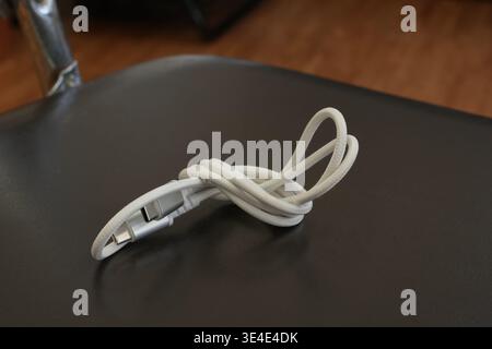 Coiled white cable rests on dark surface. Metal plug gleams subtly at one end. Background shows wooden floor and metallic object. Simple, clean compos Stock Photo
