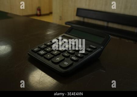 Black calculator rests on dark wooden table. Soft light highlights its buttons and screen. Background shows a chair and wall in muted tones. Still obj Stock Photo
