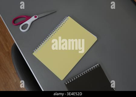 Yellow notepad rests on gray desk. Pink-handled scissors lie beside it. Black notebook sits nearby, partially open. Modern workspace, minimalist setup Stock Photo