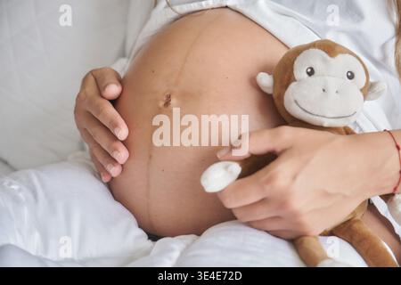 Pregnant woman embracing her baby bump while holding a soft monkey plush toy Stock Photo