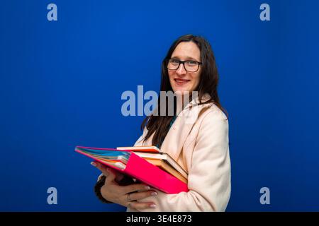 Woman wearing glasses holding a stack of colorful binders and files, smiling confidently on a blue background Stock Photo