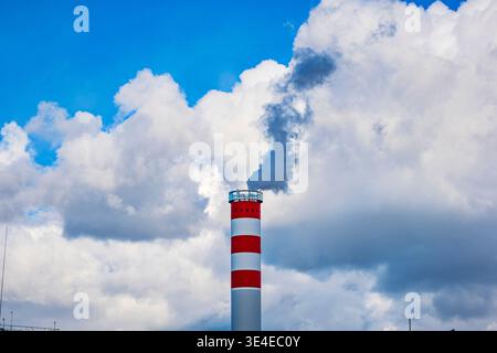 Red and white striped industrial chimney emitting dark smoke into a cloudy blue sky background Stock Photo