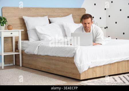 Young man in bathrobe using laptop on bed at home Stock Photo