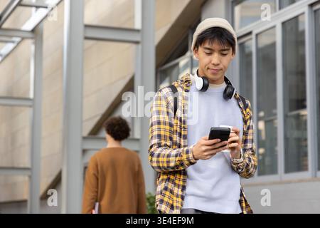 Asian man standing on walkway wearing beanie headset using phone holding cup, copy space Stock Photo