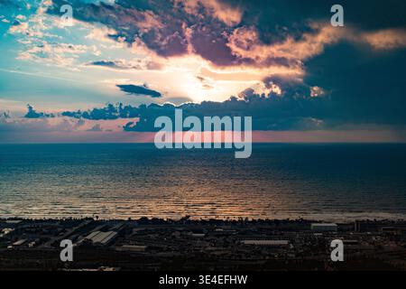 An elevated panoramic view from Mount Carmel, Haifa, Israel, looking over the Mediterranean Sea at sunset. The sun, partially hidden by large, dark, d Stock Photo