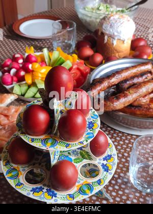Easter lunch with dyed red eggs, sausages, cucumbers, tomatoes, radishes, sandwich on plates on a stand Stock Photo