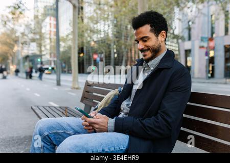 Young man checking smartphone while sitting on park bench in city Stock Photo