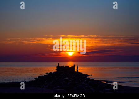 Vivid sunset over calm sea with glowing sky and rocky pier silhouette leading toward the horizon. Stock Photo