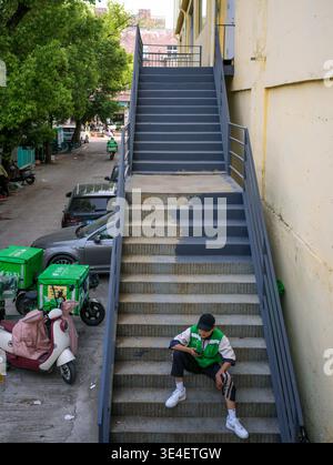 Wuhan, China - April 13, 2025: Delivery worker wearing a green uniform sitting on an external metal staircase and looking at a smartphone. The image c Stock Photo