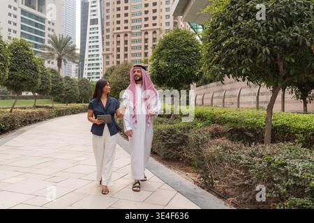 An Arabic businessman and an Asian businesswoman engage in a meeting while walking in a modern city. Stock Photo