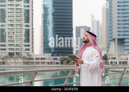 Arabic businessman in traditional attire stands on a Dubai bridge, eyeing finance and real estate opportunities in the skyline. Stock Photo