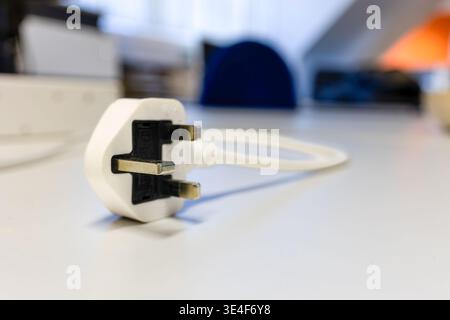 Standard British BS1363 three-pin mains electricity plug lying on grey office workstation surface. Close-up, blurred office background with natural li Stock Photo