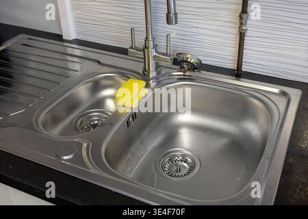 Overhead view of modern inset stainless steel kitchen sink unit. Features two bowls, a drainer area, a chrome mixer faucet, a yellow sponge, and visib Stock Photo