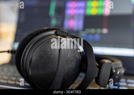 Detailed shot of professional over-ear headphones resting on laptop keyboard. Blurred screen shows digital audio workstation DAW interface with colorf Stock Photo