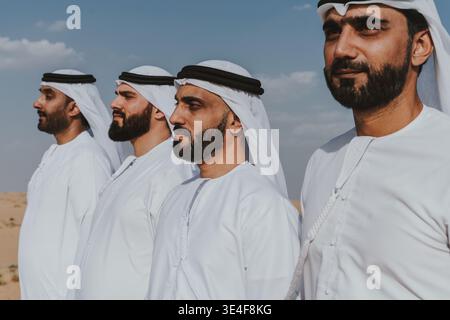 Four middle eastern friends are standing together in white dishdashas and ghutras, sharing a moment of connection in the sunny dubai desert, represent Stock Photo