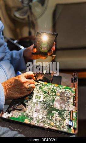 Elderly woman using smartphone flashlight for DIY laptop motherboard repair at home promoting right to repair and circular economy sustainability. Stock Photo