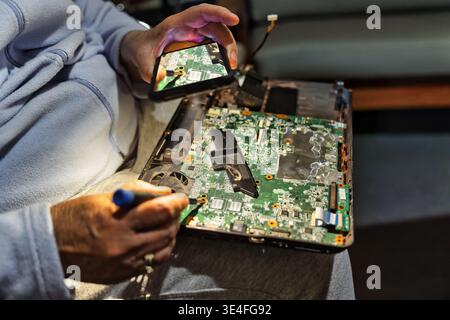 Clever senior woman using a smartphone as a magnifying glass to see tiny components while repairing a laptop at home, promoting the right to repair. Stock Photo