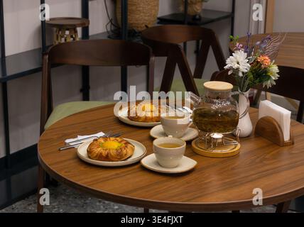Two flower-shaped pastries with fresh peach and custard cream, poured herbal tea cups and glass pot on a wooden café table with flower vase. Stock Photo