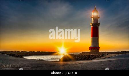 sunset view over the Atlantic Ocean in Swakopmund, featuring the historic red and white lighthouse with an illuminated beacon overlooking the rocky co Stock Photo