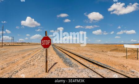 single-track railway line extends into the horizon through a barren desert landscape with a prominent red stop sign on a wooden post under a blue sky Stock Photo