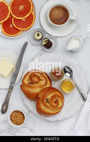 Food, Two buns with raisins, jam and honey in a white plate on a marble background for breakfast. Coffee, milk, butter, peanut butter, chocolates and Stock Photo