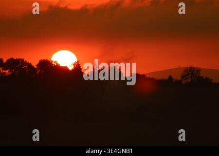 Intense red-orange sunset with the sun partially hidden behind trees and houses. Silhouetted landscape with glowing sky and atmospheric evening light. Stock Photo