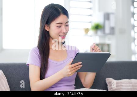 Happy Asian adult woman using a tablet and stylus. She is comfortably seated on a sofa at home, engaged in digital activity. Stock Photo