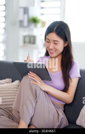 Smiling young Asian female creating or studying on a smart device with pen while relaxing comfortably in a modern interior Stock Photo