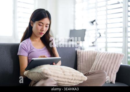 Focused Asian adult woman engaged with tablet at home Modern living room setting shows relaxed remote work or creative study Stock Photo