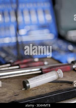 A disposable syringe lies on a workshop surface in sharp focus, with tools and an open socket set blurred in the background, 2026 Stock Photo