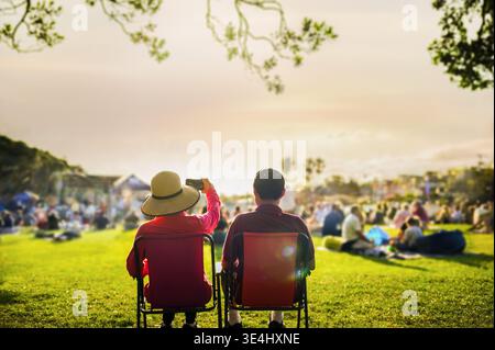 Couple taking photos using smartphone at sunset, unrecognizable crowd of people listen to the music concert in the park. Stock Photo