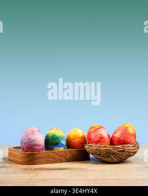 Colorful Easter eggs in a wooden egg stand and a decorative nest of sisal on a blue green gradient background with a place for text. Happy Easter concept. Greeting card. Stock Photo