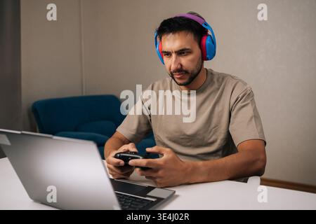 Portrait of serious focused young man wearing headphones and holding gamepad, intensely playing video game on laptop, concentrating on screen during Stock Photo
