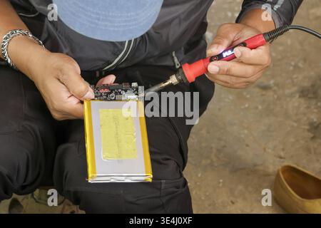 Focused technician hand repairs electronics using soldering iron on circuit board connected to flat battery during careful maintenance work requiring Stock Photo