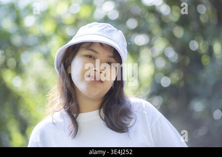 Portrait of young asian woman wearing white bucket hat and shirt outdoor posing with calm relaxed emotional expression against blurred green foliage b Stock Photo