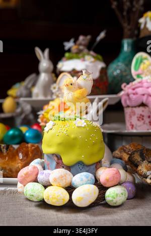 Vibrant Easter setup: green kulich topped with decorative chick and floral sprinkles, wreath of painted eggs, porcelain bunnies, decorated jars, and p Stock Photo