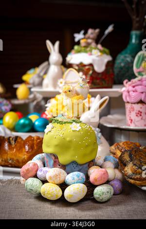 Vibrant Easter setup: green kulich topped with decorative chick and floral sprinkles, wreath of painted eggs, porcelain bunnies, decorated jars, and p Stock Photo
