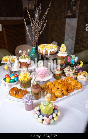 Luxurious Easter table setting on white tablecloth two-tier kulich with floral icing, egg-topped roll, wreath of painted eggs, ceramic bunnies, pastri Stock Photo