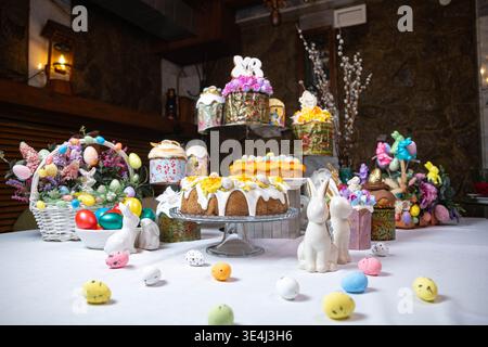 Luxurious Easter setup two-tier kulich with floral icing, basket of colorful eggs and fresh flowers, ceramic bunnies, decorated jars, and willow branc Stock Photo