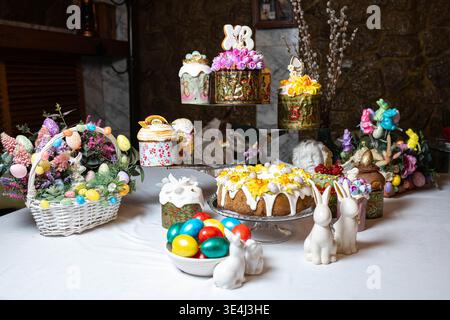 Beautifully arranged Easter table featuring various decorated kulichi on stands, white ceramic bunnies, colorful Easter eggs, woven basket with flower Stock Photo