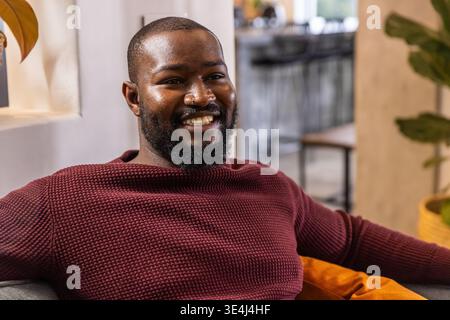 African American man in his 30s sitting on gray sofa in cafe in maroon sweater smiling Stock Photo