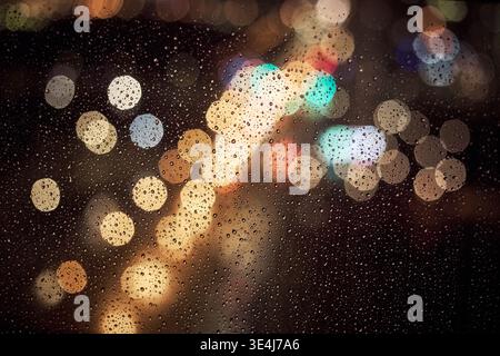 Defocused city lights seen through a rain covered window, with colorful bokeh circles, water droplets, and soft motion blur creating a moody night urb Stock Photo