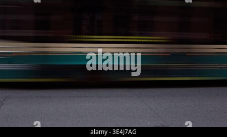 Motion blur captures a fast tram streaking past on an urban street at dusk, with dark background and textured asphalt foreground, conveying speed, com Stock Photo